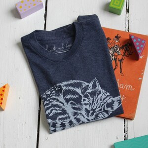 Puede incluir: Una camiseta azul marino con un gr&aacute;fico blanco de un gato durmiendo. La camiseta tiene una peque&ntilde;a etiqueta blanca con el texto "earth and hand" en ella.