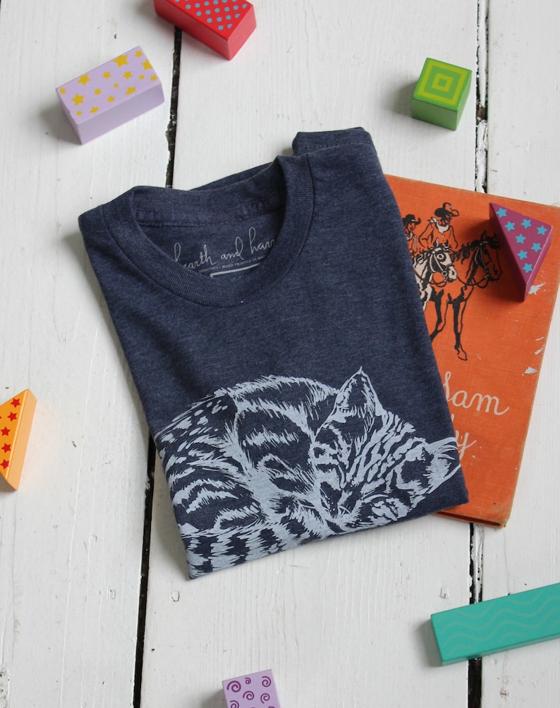 Puede incluir: Una camiseta azul marino con un gr&aacute;fico blanco de un gato durmiendo. La camiseta tiene una peque&ntilde;a etiqueta blanca con el texto "earth and hand" en ella.