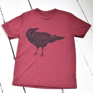 Pode incluir: Camiseta bord&ocirc; com uma ilustra&ccedil;&atilde;o preta de um corvo. O corvo &eacute; desenhado de forma detalhada e estilizada, com asas e penas vis&iacute;veis. A camiseta &eacute; feita de um material macio e &eacute; exibida em uma superf&iacute;cie de madeira branca.