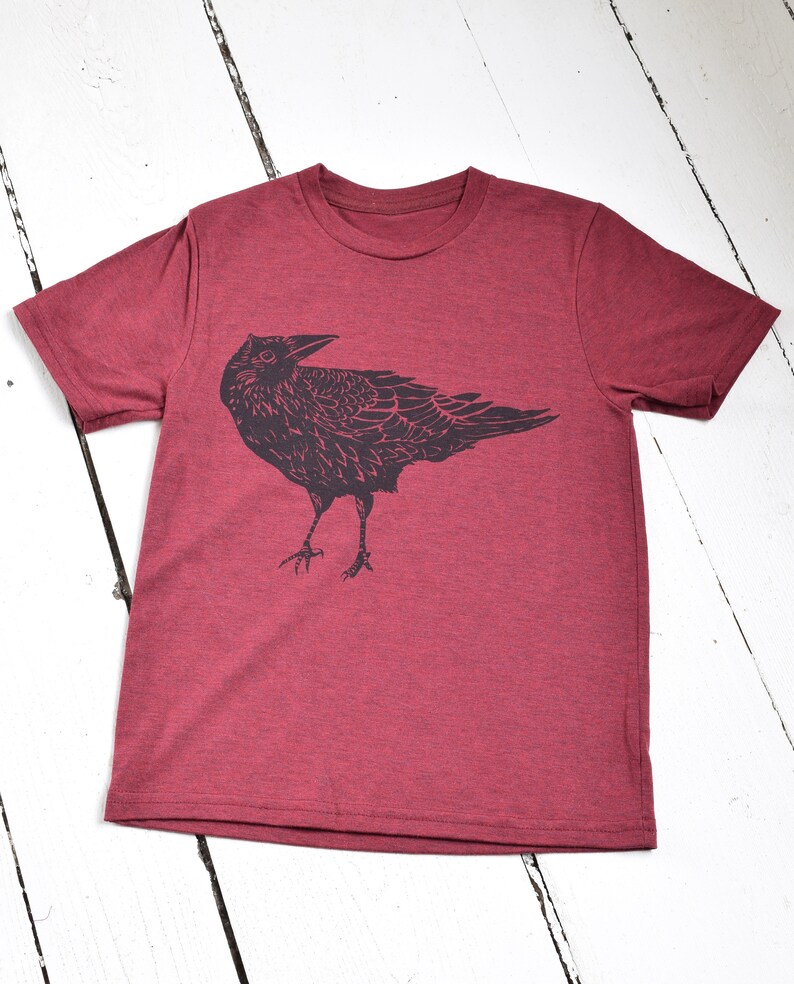 Pode incluir: Camiseta bord&ocirc; com uma ilustra&ccedil;&atilde;o preta de um corvo. O corvo &eacute; desenhado de forma detalhada e estilizada, com asas e penas vis&iacute;veis. A camiseta &eacute; feita de um material macio e &eacute; exibida em uma superf&iacute;cie de madeira branca.