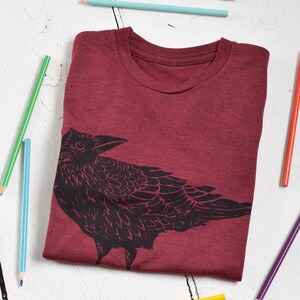 Pode incluir: Uma camiseta bord&ocirc; com gola redonda e um desenho de corvo preto. A camisa dobrada &eacute; cercada por l&aacute;pis coloridos em uma superf&iacute;cie de madeira branca. O desenho do corvo &eacute; uma silhueta detalhada.