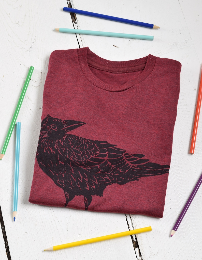 Pode incluir: Uma camiseta bord&ocirc; com gola redonda e um desenho de corvo preto. A camisa dobrada &eacute; cercada por l&aacute;pis coloridos em uma superf&iacute;cie de madeira branca. O desenho do corvo &eacute; uma silhueta detalhada.