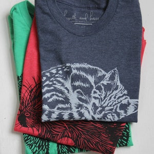 Puede incluir: Tres camisetas dobladas con diferentes dise&ntilde;os de animales. La camiseta de arriba es azul marino con un dise&ntilde;o de gato blanco y gris. La camiseta del medio es roja con un dise&ntilde;o de puercoesp&iacute;n negro y rojo. La camiseta de abajo es verde con un dise&ntilde;o de puercoesp&iacute;n negro y verde. Las camisetas tienen el texto "earth and hand" impreso en la camiseta de arriba.