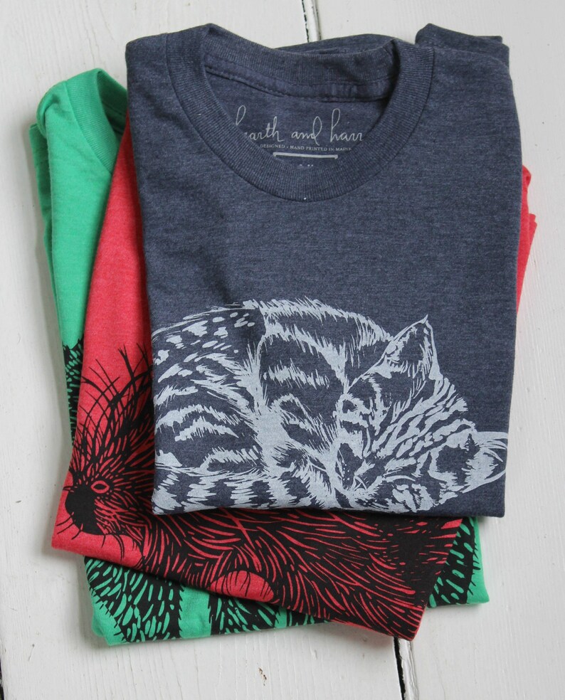 Puede incluir: Tres camisetas dobladas con diferentes dise&ntilde;os de animales. La camiseta de arriba es azul marino con un dise&ntilde;o de gato blanco y gris. La camiseta del medio es roja con un dise&ntilde;o de puercoesp&iacute;n negro y rojo. La camiseta de abajo es verde con un dise&ntilde;o de puercoesp&iacute;n negro y verde. Las camisetas tienen el texto "earth and hand" impreso en la camiseta de arriba.