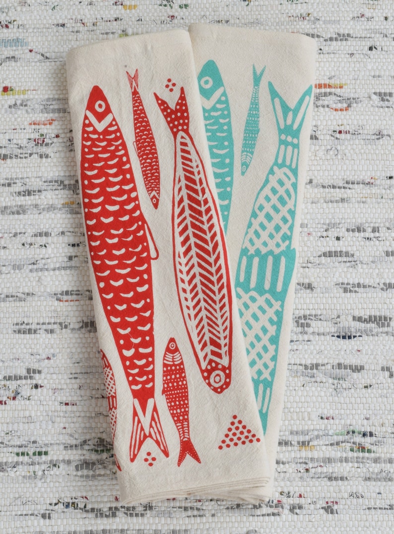 May include: Two white kitchen towels with red and blue fish designs. The fish are stylized and have a folk art feel. The towels are folded and displayed on a white background.