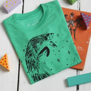 Puede incluir: Una camiseta verde con una ilustraci&oacute;n en blanco y negro de un zorro saltando frente a un cielo estrellado. El texto "with and ho" est&aacute; impreso en la camiseta.