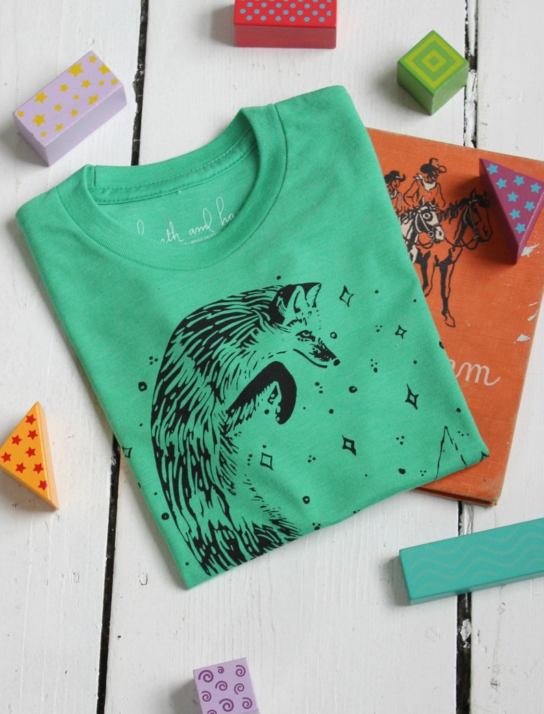 Puede incluir: Una camiseta verde con una ilustraci&oacute;n en blanco y negro de un zorro saltando frente a un cielo estrellado. El texto "with and ho" est&aacute; impreso en la camiseta.