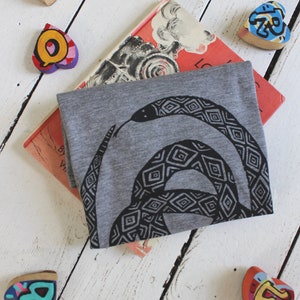 May include: A gray t-shirt with a black and white graphic print of a snake with a geometric pattern. The shirt is folded and lying on a white wooden surface with colorful wooden hearts scattered around it. The shirt is partially covering a book with the title "Locomotive" visible.