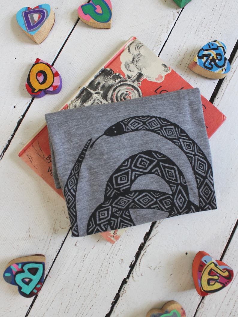May include: A gray t-shirt with a black and white graphic print of a snake with a geometric pattern. The shirt is folded and lying on a white wooden surface with colorful wooden hearts scattered around it. The shirt is partially covering a book with the title "Locomotive" visible.