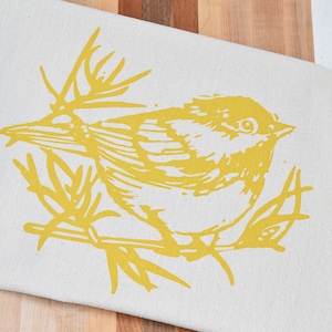 May include: A yellow bird with a detailed design is printed on a beige fabric kitchen towel. The towel is folded over a wooden cutting board with multiple wood tones.