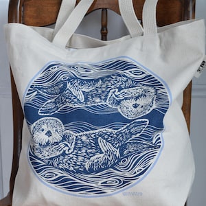 May include: Cream-colored tote bag featuring a blue circular design with two otters floating in water. The otters are depicted in a detailed linocut style. The bag has cream-colored handles and is sitting on a wooden chair.