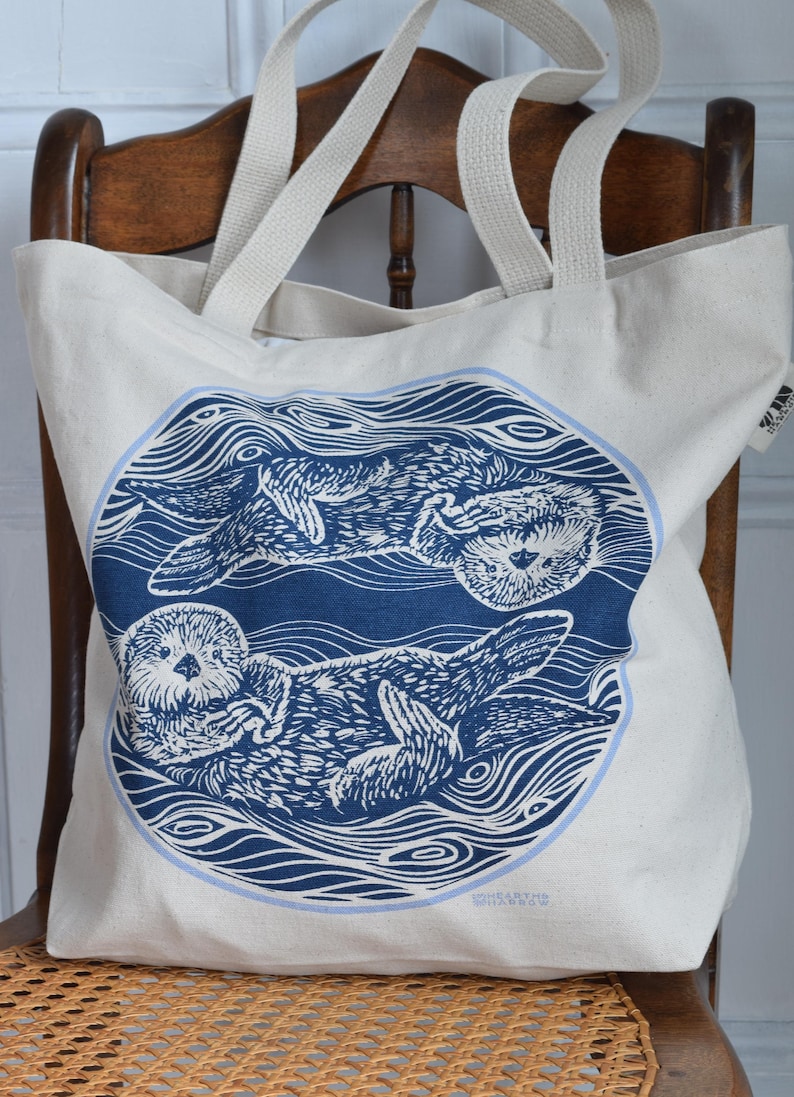 May include: Cream-colored tote bag featuring a blue circular design with two otters floating in water. The otters are depicted in a detailed linocut style. The bag has cream-colored handles and is sitting on a wooden chair.