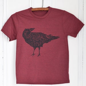 Pode incluir: Camiseta bord&ocirc; com um gr&aacute;fico preto de um corvo. O corvo &eacute; retratado em uma ilustra&ccedil;&atilde;o estilizada e detalhada. A camiseta est&aacute; pendurada em um cabide de madeira em um fundo branco.