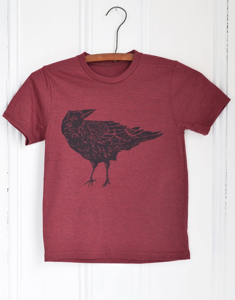 Pode incluir: Camiseta bord&ocirc; com um gr&aacute;fico preto de um corvo. O corvo &eacute; retratado em uma ilustra&ccedil;&atilde;o estilizada e detalhada. A camiseta est&aacute; pendurada em um cabide de madeira em um fundo branco.