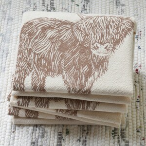 May include: A stack of five cream-colored tea towels with a brown Highland cow design printed on them.