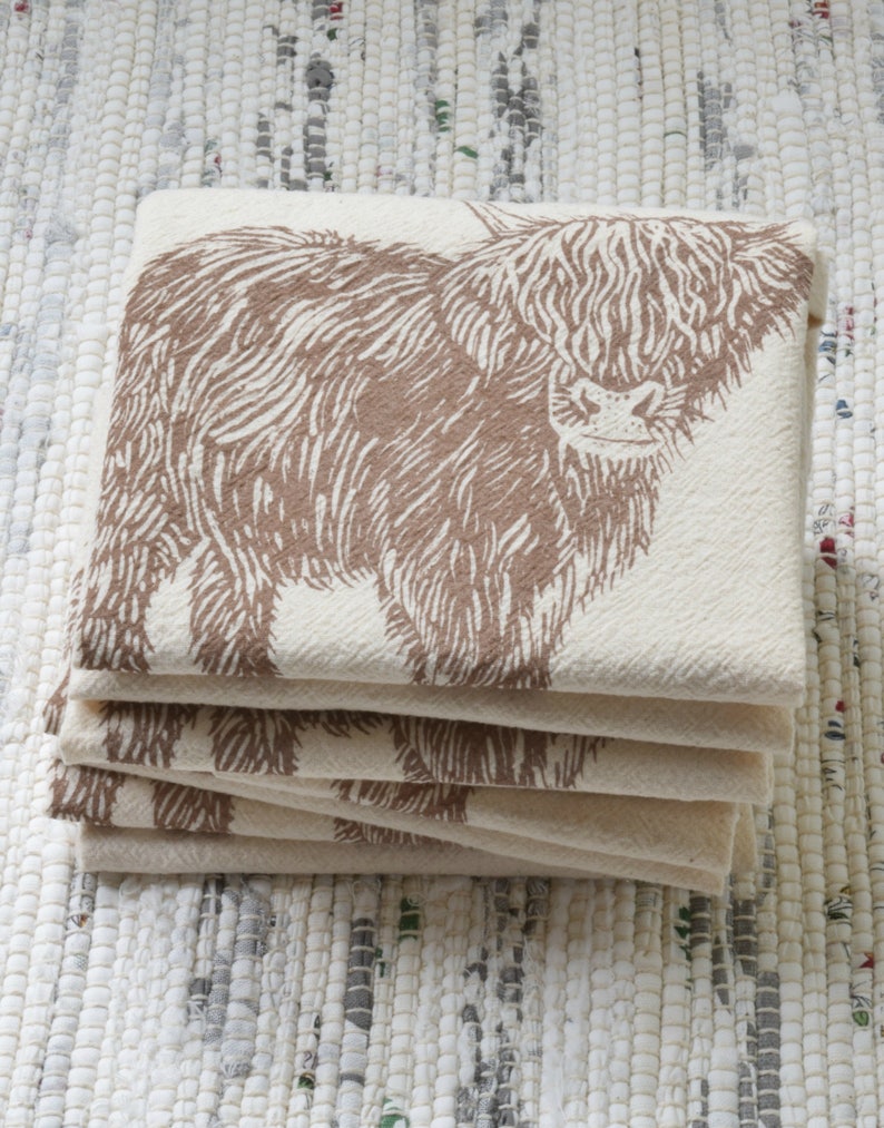 May include: A stack of five cream-colored tea towels with a brown Highland cow design printed on them.