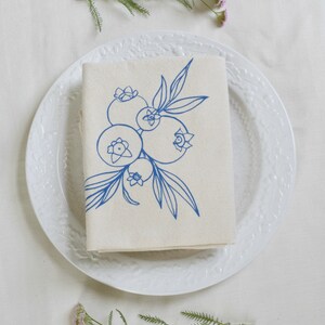 May include: A folded beige linen napkin with a blue blueberry illustration, placed on a white plate. The plate is surrounded by sprigs of pink flowers and greenery, creating a fresh, natural aesthetic.