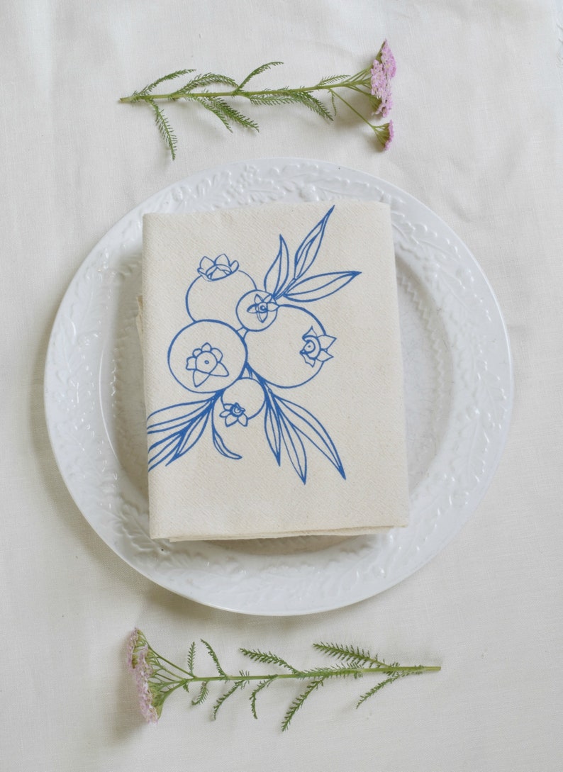 May include: A folded beige linen napkin with a blue blueberry illustration, placed on a white plate. The plate is surrounded by sprigs of pink flowers and greenery, creating a fresh, natural aesthetic.