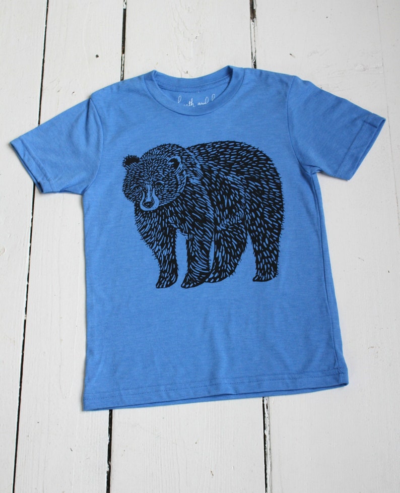 May include: A blue t-shirt with a black and white illustration of a bear on the front. The text "with and" is visible on the collar.