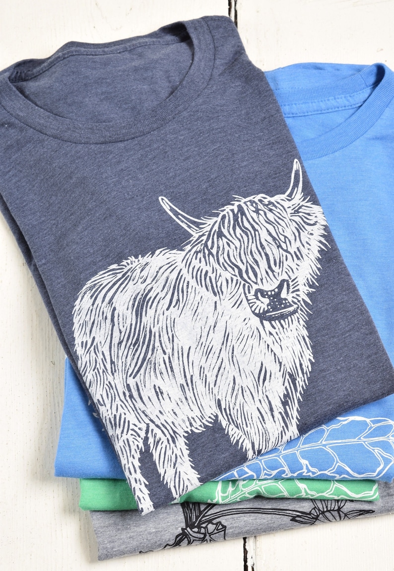 May include: A stack of four t-shirts with different designs. The top shirt is a dark blue heather color with a white graphic of a Highland cow. The second shirt is a light blue color. The third shirt is a light green color. The bottom shirt is a gray heather color.