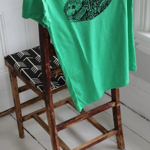 K&ouml;nnte beinhalten: Ein leuchtend gr&uuml;nes T-Shirt mit einer schwarzen Illustration eines Seeotters liegt &uuml;ber der R&uuml;ckenlehne eines Holzstuhls. Der Stuhl hat einen Sitz mit einem schwarz-wei&szlig;en Pfeilmuster. Der Stuhl und das T-Shirt befinden sich in einem Raum mit wei&szlig;en W&auml;nden und einem wei&szlig;en Boden.