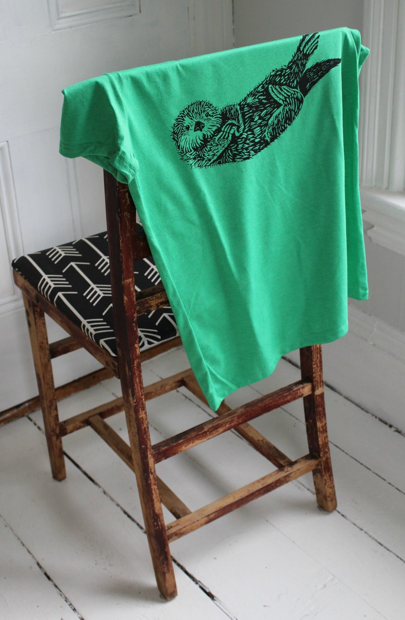 K&ouml;nnte beinhalten: Ein leuchtend gr&uuml;nes T-Shirt mit einer schwarzen Illustration eines Seeotters liegt &uuml;ber der R&uuml;ckenlehne eines Holzstuhls. Der Stuhl hat einen Sitz mit einem schwarz-wei&szlig;en Pfeilmuster. Der Stuhl und das T-Shirt befinden sich in einem Raum mit wei&szlig;en W&auml;nden und einem wei&szlig;en Boden.