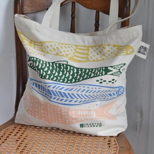 May include: A cream-colored tote bag with four colorful fish designs in yellow, green, blue, and peach. The bag has two cream-colored handles and is sitting on a wooden chair. The brand name "HEARTH & HARROW" is printed on the bottom.