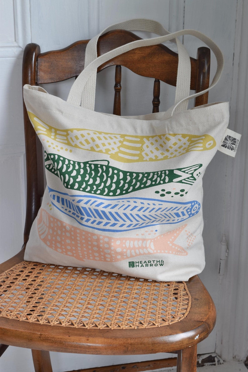 May include: A cream-colored tote bag with four colorful fish designs in yellow, green, blue, and peach. The bag has two cream-colored handles and is sitting on a wooden chair. The brand name "HEARTH & HARROW" is printed on the bottom.