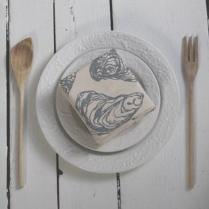May include: A white dinner plate with a decorative border, a smaller white plate inside, and a folded linen napkin with a blue oyster print on top.