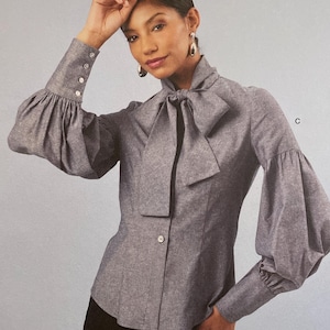 May include: Simplicity sewing pattern S3032 featuring a woman wearing a gray blouse with a bow and puffed sleeves. The pattern includes size information in U.S. sizes 18-26. The image also shows illustrations of the blouse in green and blue.