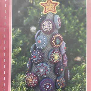 May include: A pink package featuring a photo of a handmade Christmas tree. The tree is adorned with colorful, embroidered circular ornaments and topped with a gold star. The text reads "Macaroon Christmas Tree by Reiko Kato" and "Stitch Publications."