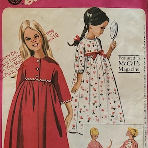 May include: A vintage McCall's sewing pattern for a girl's nightgown and robe with a matching Barbie doll costume. The pattern is printed on a white background with pink and red accents. The pattern number is 7545 and the price is 50 cents. The pattern features a girl wearing a red nightgown and a girl wearing a pink robe. The pattern also includes a doll wearing a pink nightgown and a doll wearing a pink robe. The pattern is from 1964 and is licensed by Mattel.