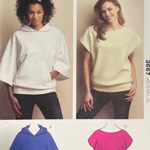May include: A Kwik Sew 3667 sewing pattern envelope for misses' sizes XS-XL. The front displays two garment styles: a white hooded top with a front pocket and a cream-colored short-sleeved top. Illustrations of a blue hoodie and a pink top are included.