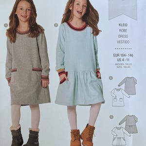 May include: A Burda Kids sewing pattern envelope 9286 featuring two dress designs. View A is a grey long-sleeve dress with pockets. View B is a light blue long-sleeve dress with a gathered skirt. The package includes sizes EUR 104-146 and US 4-11.