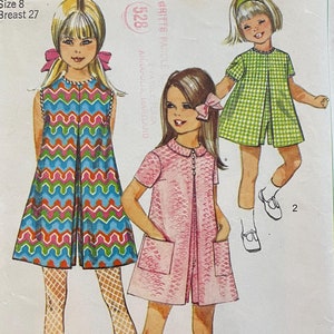 May include: A vintage Simplicity sewing pattern for a girl's dress. The pattern number is 7464 and the price is 60 cents. The pattern includes three different styles of dresses, one with a geometric print, one with a pink solid color, and one with a green gingham print. The pattern is for size 8 and requires a 27 inch bust.
