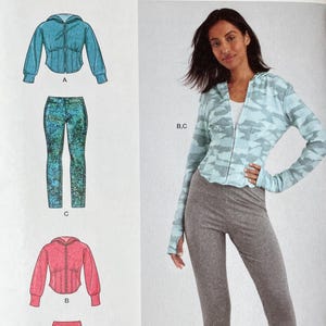 May include: Simplicity sewing pattern S9895 featuring a hooded jacket and pants. The pattern includes sizes 20W-28W. The image shows a woman wearing a light blue camouflage print jacket and gray pants. The pattern is designed and printed in the USA.