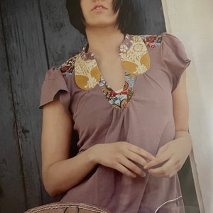 May include: A sewing pattern for a women's tee shirt called "Mariposa Tee" by Sew Liberated. The pattern is number 112. The image shows a model wearing a purple tee shirt with a colorful floral collar.