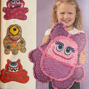 May include: Simplicity pattern 1237 for making monster-shaped pillows. The pattern features various monster designs in pink, red, and orange. A young girl holds a finished purple monster pillow with a smiling face. The pattern is one size.