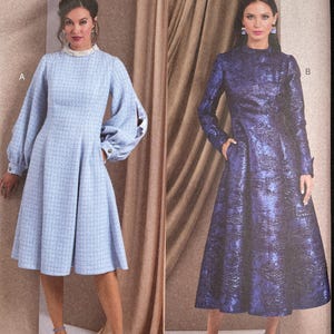 May include: Vogue Patterns V1983. The image shows two women modeling dresses. Dress A is light blue with long sleeves and a flared skirt. Dress B is a dark blue, textured dress with long sleeves and a flared skirt. The Vogue Patterns logo is at the top.