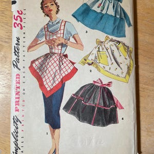 May include: Vintage Simplicity pattern envelope featuring apron designs. The cover displays a woman in a blue top and skirt, wearing a red and white checkered apron. Three additional apron styles are illustrated: striped, floral, and black with pink ribbons. The text reads "ONE-YARD APRONS".