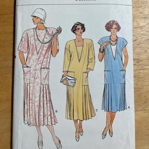 May include: Vogue Patterns sewing pattern envelope featuring three dress designs. The dresses are in pink, yellow, and blue, each with a V-neck and pleated skirt. The envelope has the Vogue logo and pattern details.