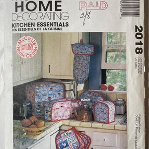 May include: A McCall's Home Decorating sewing pattern envelope for kitchen essentials, featuring illustrations of fabric covers for appliances, a hanging bag, and quilted pot holders in blue, red, and white patterns with a kitchen scene background.