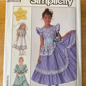 May include: Simplicity sewing pattern for a girl's dress. The pattern includes three different styles: a white dress with lace, a green floral dress, and a purple dress with lace trim. The pattern is size 8. The pattern is titled "Made in Heaven".