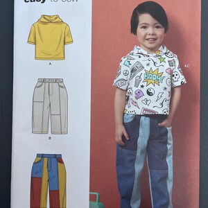May include: Simplicity sewing pattern R12145 for children's clothing. The pattern includes instructions for a short-sleeved hoodie and various styles of pants. The image shows a child wearing the finished outfit, featuring a white hoodie with colorful graphics and patchwork jeans.