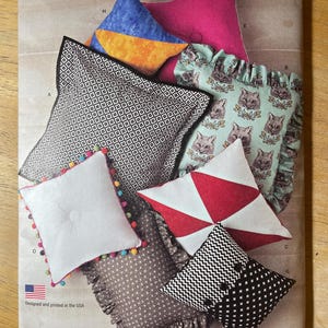 May include: Simplicity pattern featuring various pillow designs. Includes pillows with geometric patterns, floral prints, and solid colors. Some pillows have ruffles or pom-pom trim. The pattern is designed and printed in the USA.