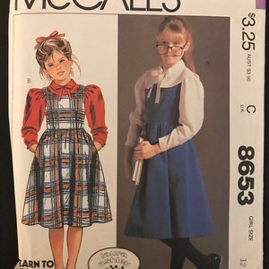 May include: McCall's sewing pattern for a girl's dress, pattern number 8653, size 12. The pattern shows two different styles of dresses, one with a plaid skirt and red sleeves, and the other with a solid blue skirt. The pattern is for a girl's dress.