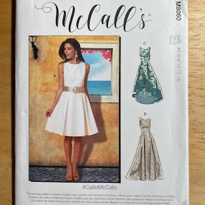 May include: McCall's sewing pattern featuring multiple dress designs. The cover shows a woman in a white dress with a gold belt. The pattern includes the text "#CallieMcCalls" and is designed and printed in the USA.