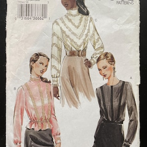 May include: A Vogue Patterns sewing pattern envelope, number 7734, featuring illustrations of three long-sleeved blouses with lace trim details. The designs include a high-neck cream blouse, a pink blouse with vertical lace, and a black blouse with dark trim.