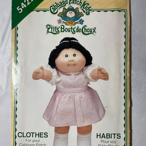 May include: A See & Sew 5422 sewing pattern envelope for Cabbage Patch Kids clothes. The cover features a doll with black yarn hair, a white blouse, and a pink and white striped pinafore dress. Text includes See & Sew, 5422, and Cabbage Patch Kids.