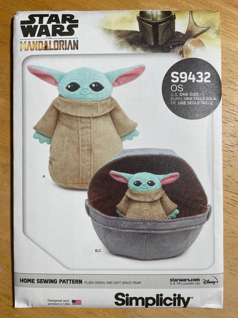 May include: A sewing pattern featuring a plush Grogu and soft space pram. The pattern includes images of the Grogu plush toy in a tan coat with large ears and a gray pram. The Star Wars Mandalorian logo is at the top.